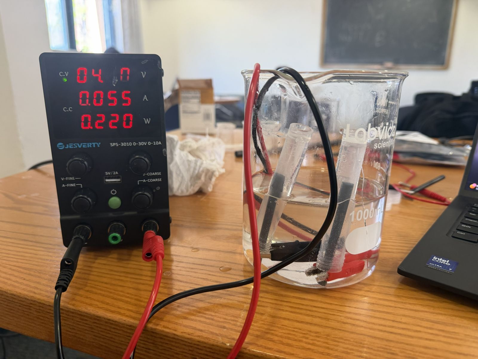 Electrolysis setup: a DC power supply reading 4.0 V wired to graphite electrodes in a beaker of electrolyte, with collection tubes over each electrode